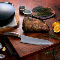 Suncraft Elegancia kokkiveitsi leikkuulaudalla, ympärillä yrttejä, sitruuna ja valkosipulia. SG2-teräs ja Damascus-pinta.