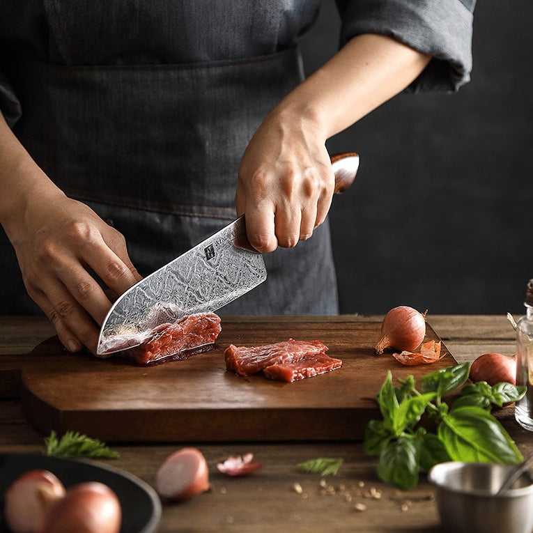Kokki käyttää XINZUO Jiang Damaskus Santokuveistä, 18 cm, leikatakseen lihaa puuleikkuulaudalla, osa premium-veitsisarjaa.
